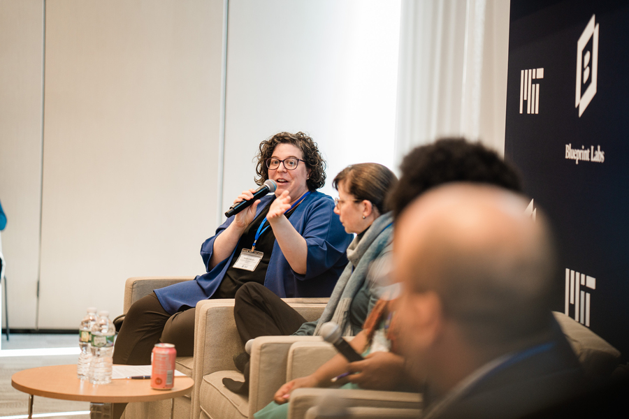 Four people sit in a row of easy chairs. The woman farthest from the camera speaks into a microphone while looking at the other panelists.