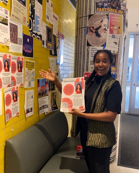 Sophia Ashebir pauses while putting up AgeLab posters on a yellow wall.