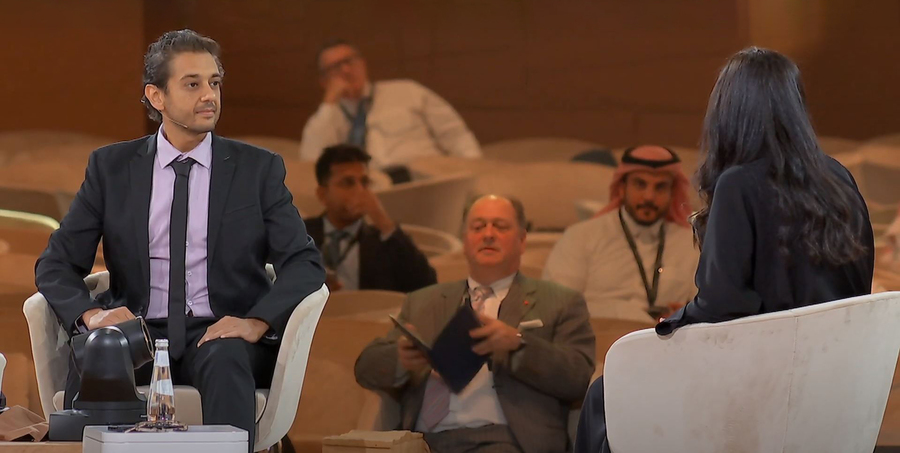 Mohammed Yahia sits on a stage. He's wearing a microphone and speaking to someone out of the frame. An out-of-focus audience is in the background. 