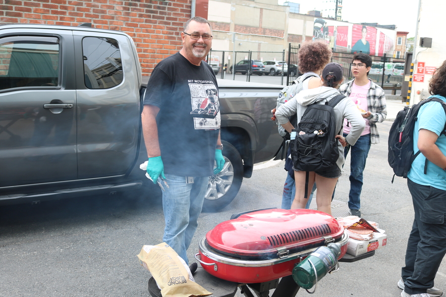 Pat McAtamney stands in front of a parked pickup truck, smiling behind a smoking grill on the street 
