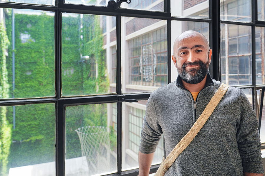 Photo of Pedro Reynolds-Cuéllar, who stands in front of a large window divided into small panes overlooking the courtyard of an ivy-covered industrial-style building.