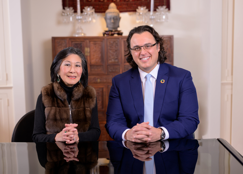 Lisa Yang and Amos Winter pose for a photo. They sit together at a black table in a white room with an intricately carved wooden dresser and a Buddah head statue in the background.