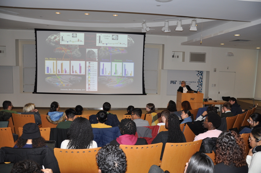 At the front of a theater-style auditorium, Nancy Kanwisher stands at a podium in front of several rows of students looking at a slides of a technical presentation.