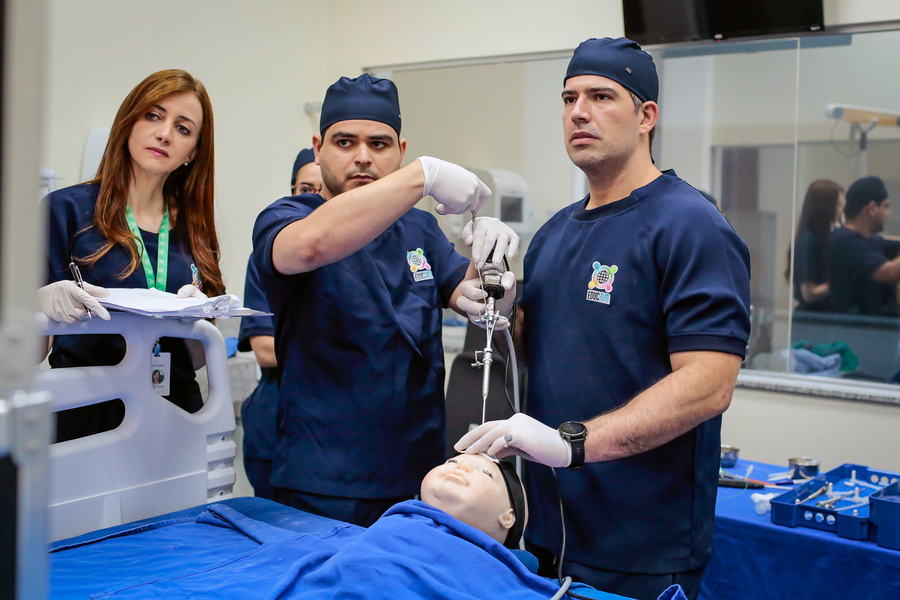 Two surgical residents in hospital garb hold medical equipment over a physical model of an infant. One holds the wires while the other guides an instrument to insert a rod into the brain of the model. At left, Giselle Coelho, also in medical garb, looks on.