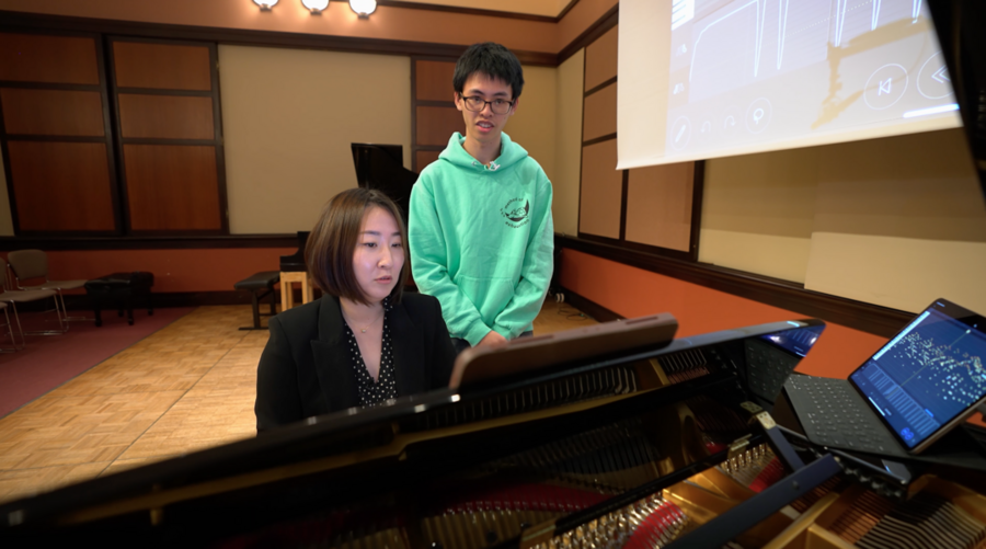 Mi-Eun Kim, seated, plays a piano while Holden Mui, standing behind her, watches. An open laptop with a visual representation of data rests atop the piano.