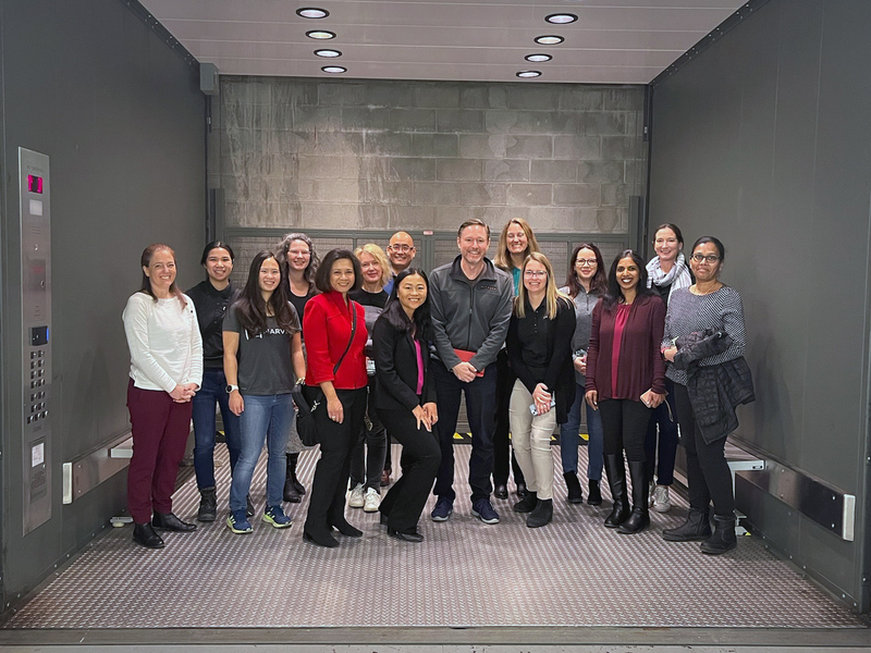 15 people pose together for a photo in a freight elevator so large that they have lots of space around them.