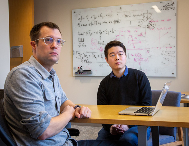 Two men sitting at a table, with a whiteboard in the background. 