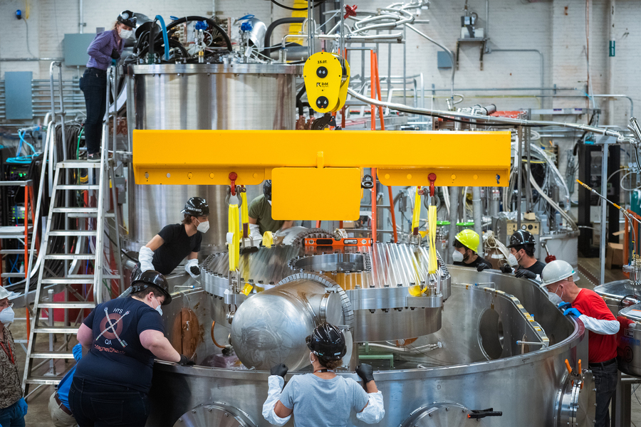 Group of seven people, while wearing hard hats and face masks, surround cylindrical container as a magnet is lowered