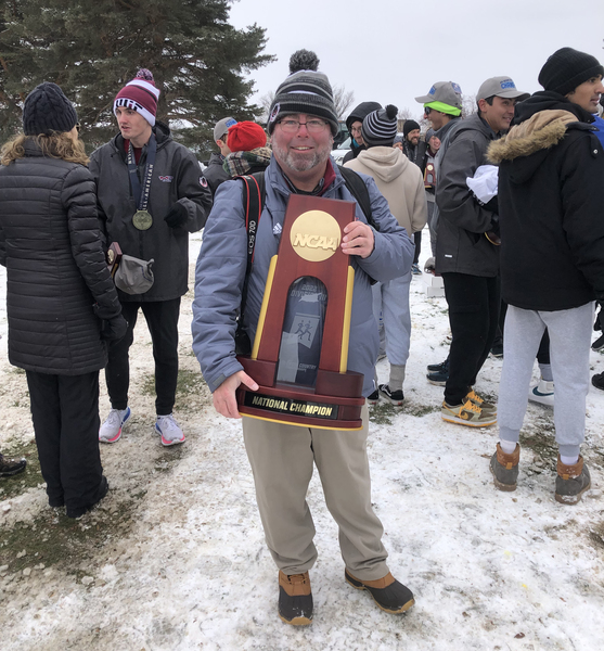 Photo of Ken Johnson outside in cold-weather gear holding a large NCAA trophy, while about a dozen others gather behind him, talking to each other