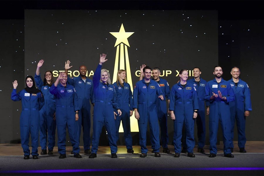 12 astronauts in blue jumpsuits appear on stage. Several are clapping or waving at the crowd.