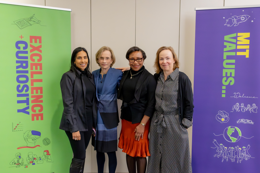 Four women pose in a line against a wall, flanked by banners reading “Excellence Curiosity” and “MIT Values…"