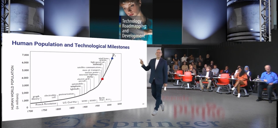 On a stage, Oli De Weck points to a large line graph plotting human tech milestones against population and time.