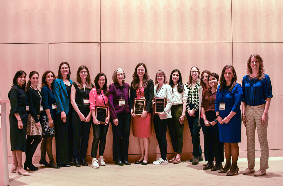 15 women stand in a semicircle. Three hold plaques. 