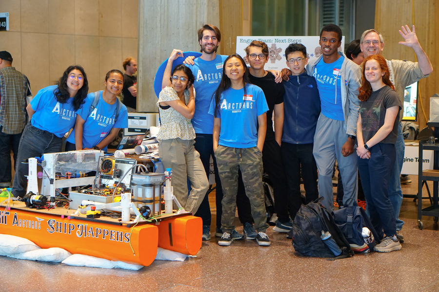 Nine students from the Arcturus team and their advisor smile and wave in their team T-shirts, behind their orange boat.