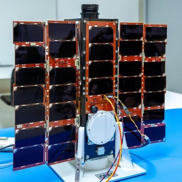 The AEROS-MH1 CubeSat sits on a tabletop. It looks like five columns of solar cells flanking a spacecraft body that's only partly visible, 