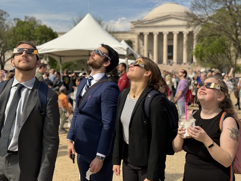 Four people gaze skyward through eclipse glasses on a sunny day; in the background, a large number of people are doing the same. The Jefferson Memorial is in the far background.