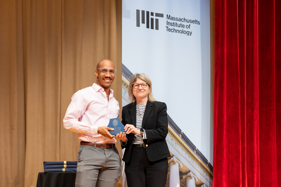 A man wearing a button-down shirt and glasses is being handed an award by a woman who is smiling and wearing a black suit.