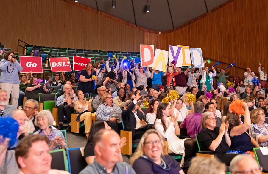 Audience shot of an auditorium full of people who are smiling, clapping, and holding signs of encouragement.