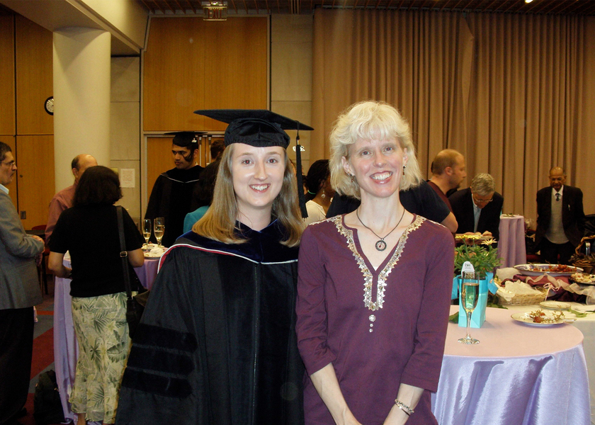Mary Ellen Wiltrout, wearing black graduation regalia, poses for a photo with Tania Baker in a reception hall.