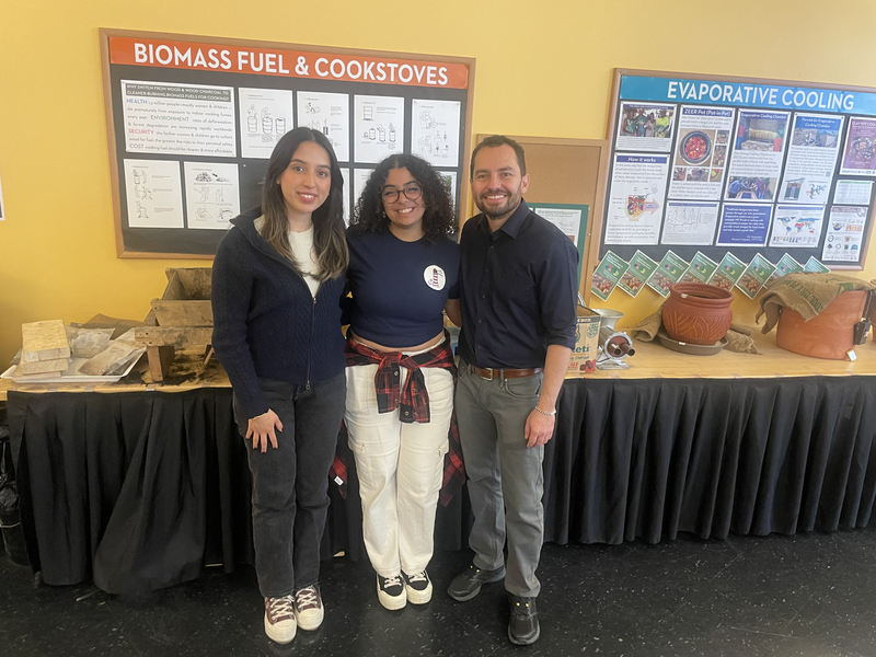 Mercy Torres, Sandra Youssef, and Rony Herrarte pose together in front of a table and posters demonstrating biomass fuel, cookstoves, and evaporative cooling. 