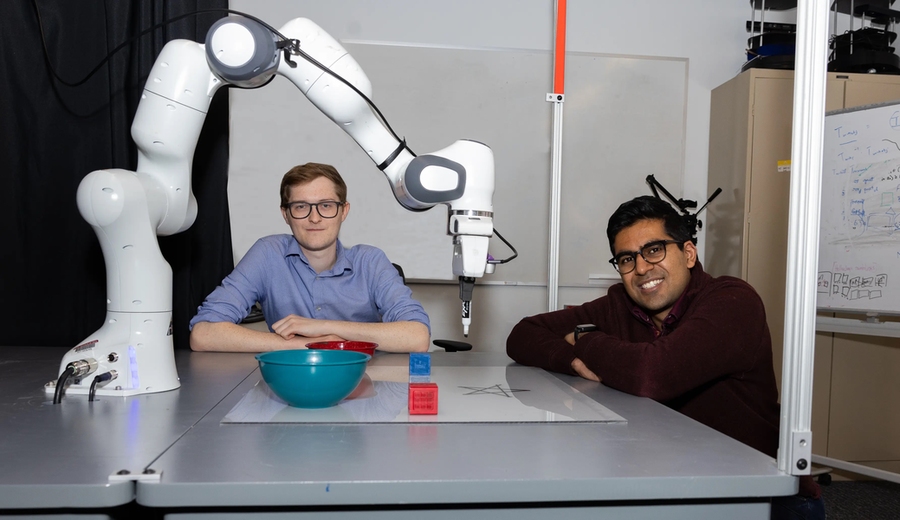 Two students sit at a table dominated by a white robotic arm that drew a star on a whiteboard.
