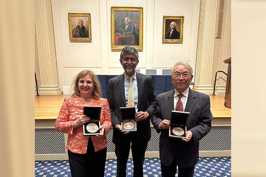 Daniela Rus, Takeo Kanade, and Vijay Kumar pose together displaying medallions held in boxes. Behind them are portraits of Ben Franklin and other American founders.