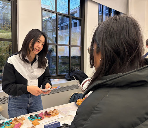 Angelina Wu, standing at a table laden with pastries, prepares to hand a brochure to someone