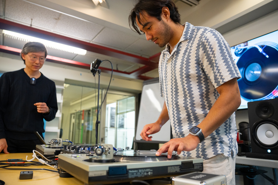 Philip Tan guides student Karl Velazquez, who is standing over a turntable with his finger on the platter