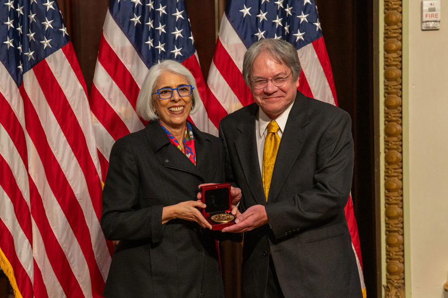 Arati Prabhakar and Larry Edwards pose together with a medal, in front of a U.S. flag