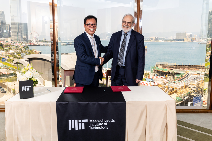 Sebastian Man and Eric Grimson shake hands while standing behind a table draped with an MIT banner, a cityscape visible through the window behind them..