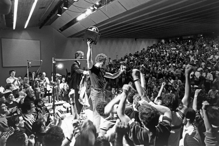 Grayscale photo of hundreds of MIT students in a large classroom in the early 1980s. A student raises a trophy as others cheer.
