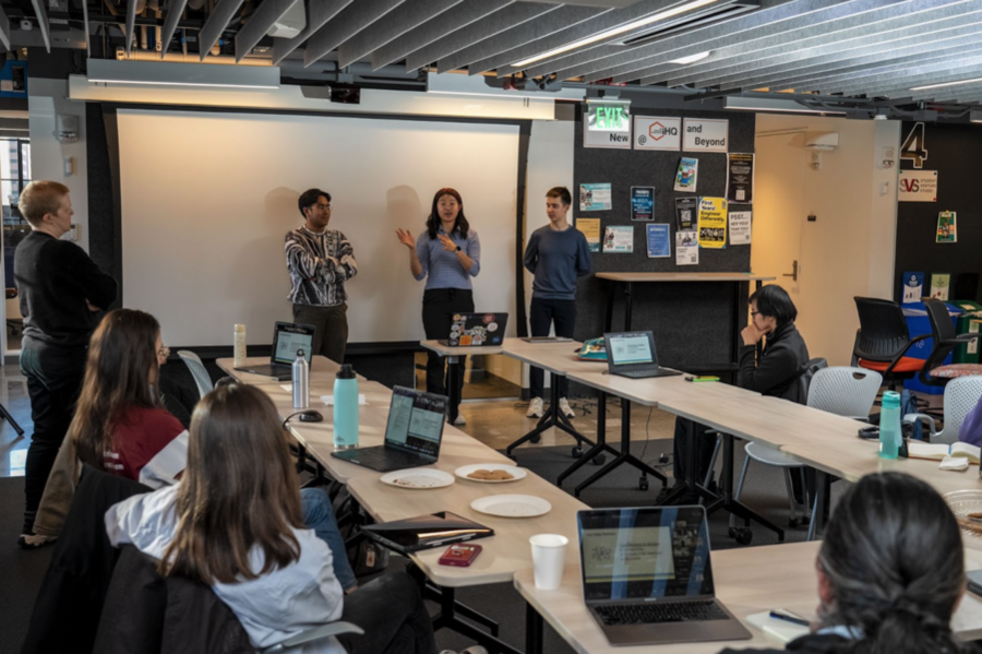 Three people stand before an unused projection screen in front of a classroom; several students with their backs to the camera sit around a U-shaped table 