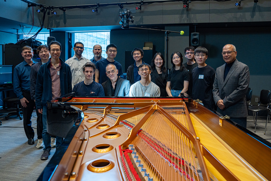 16 people in two rows pose behind an open piano case