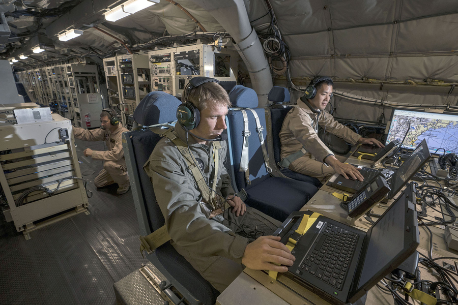 Two personnel are seated in a military test aircraft operating communications equipment while a third works in the background.
