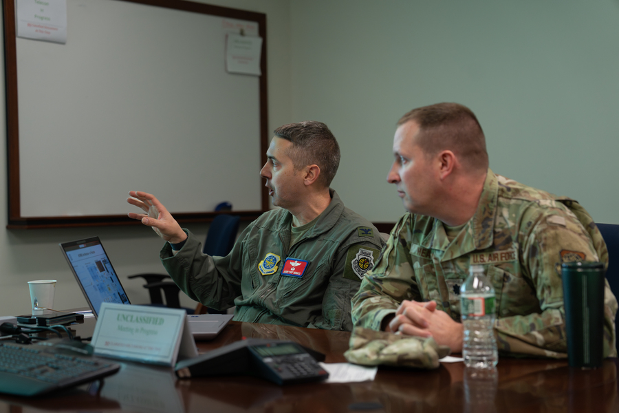 Joseph Monaco and Tim Heaton, both in military fatigues, sit at a conference table looking at something outside the field of view