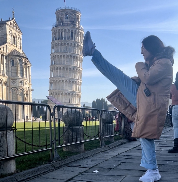 Crystal Liang poses humorously, staging a high kick to appear as though her leg is supporting the Leaning Tower of Pisa. 