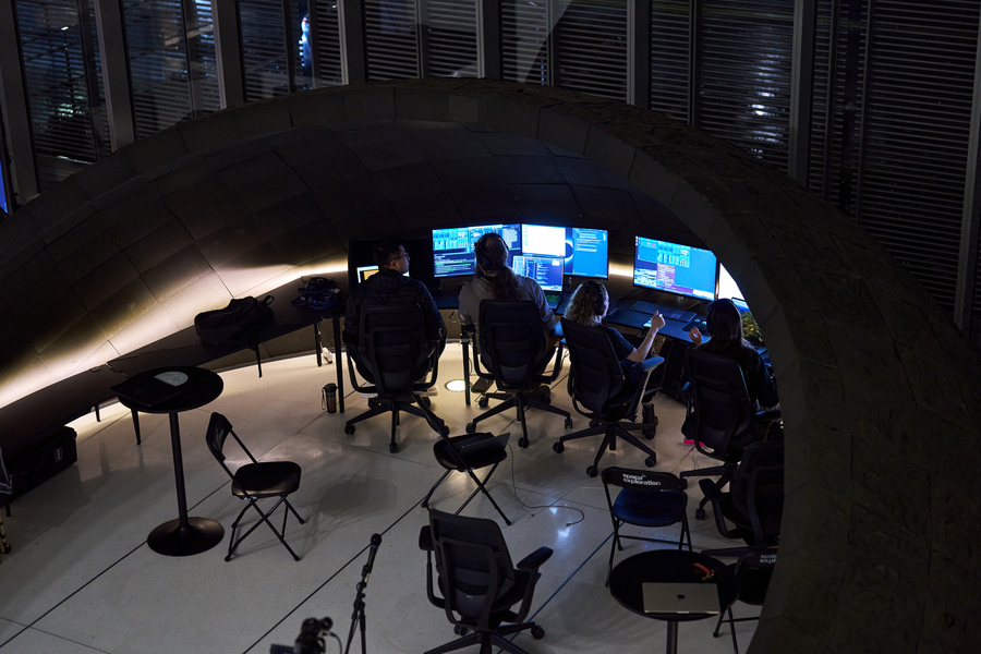 Four people sit at computer workstations under a large, curved architectural structure. 