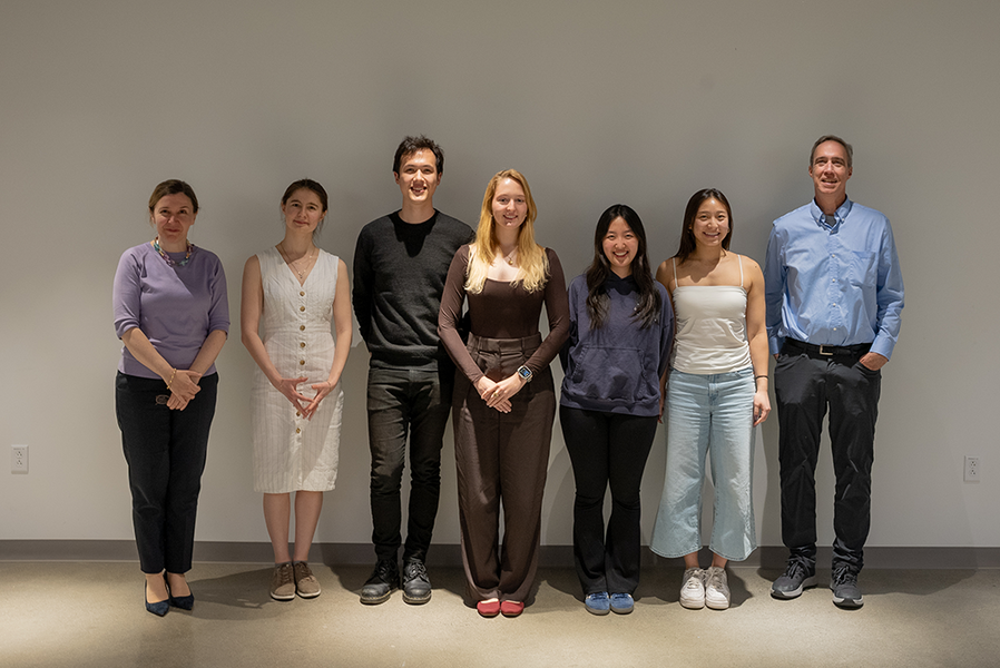 Seven smiling people pose in a line against a beige-colored wall.
