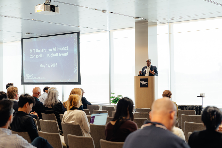 A man in a suit stands at a lectern in front of rows of people in chairs. A blue slide behind him reads MIT Generative AI Impact Consortium Kickoff Event.