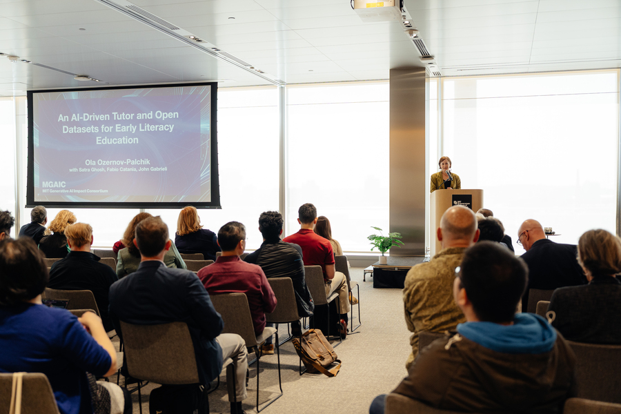 A woman with short orange hair stands at a lectern in front of rows of people in chairs. A blue slide behind her reads "AI-Driven Tutors and Open Datasets for Early Literacy Education."