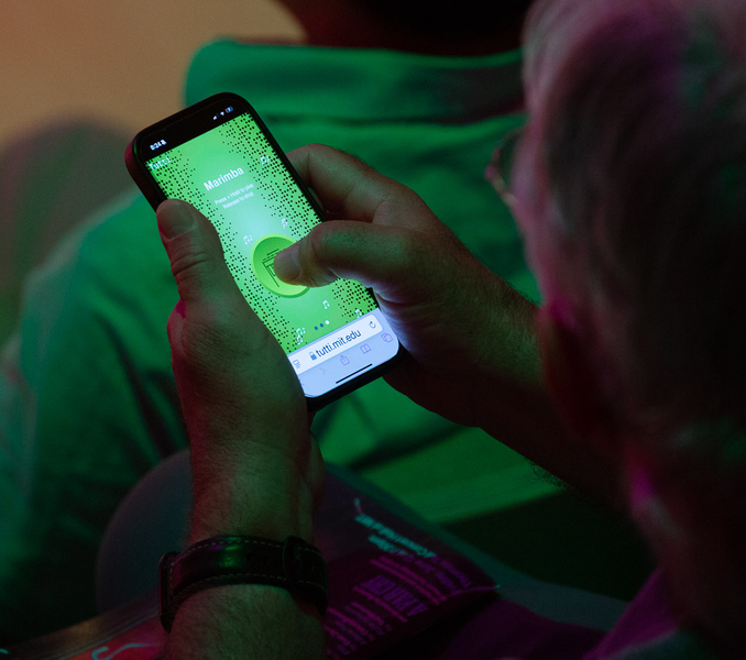 A green cellphone screen with many black dots displays the word "Marimba." The user's thumb is on a large button in the center.