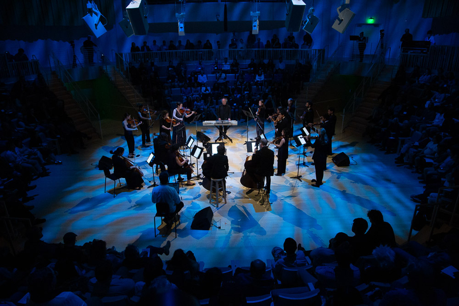 18 musicians in a circle on a blue-lit stage surrounded by audience members