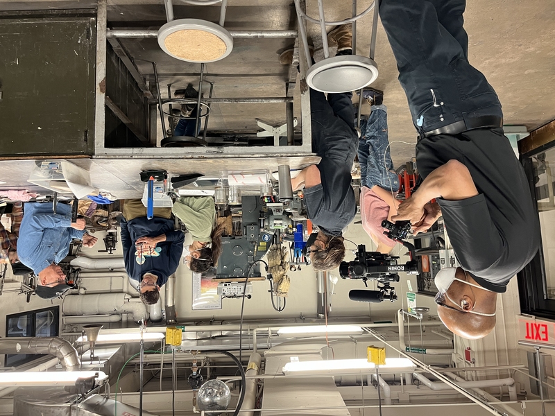 Candid photo of a cameraman filming in an industrial classroom setting where an instructor is speaking with four college students wearing protective gear around a table.