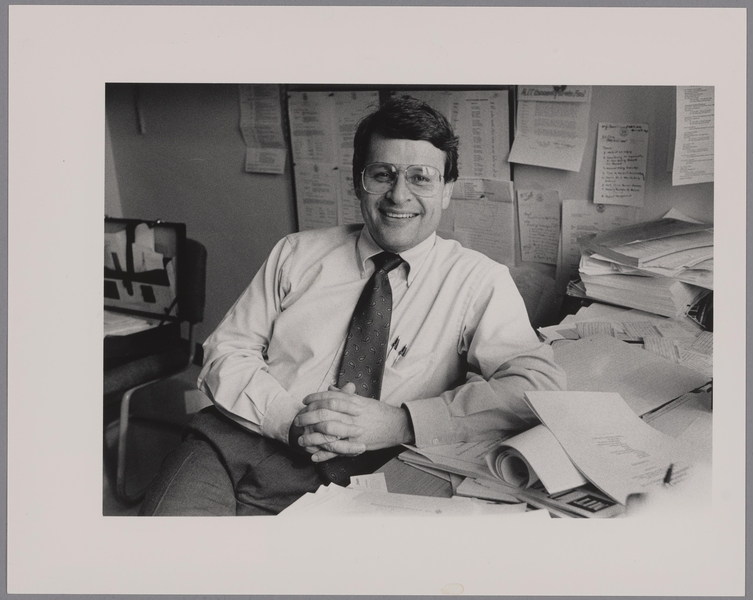Grayscale photo of a man seated at a desk buried in papers. More paper is tacked to the wall behind him.