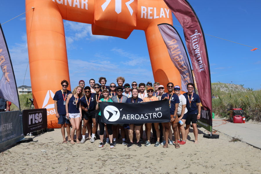 22 smiling teammates stand under a large orange inflatable arch on sandy terrain, holding an "AeroAstro MIT" banner.