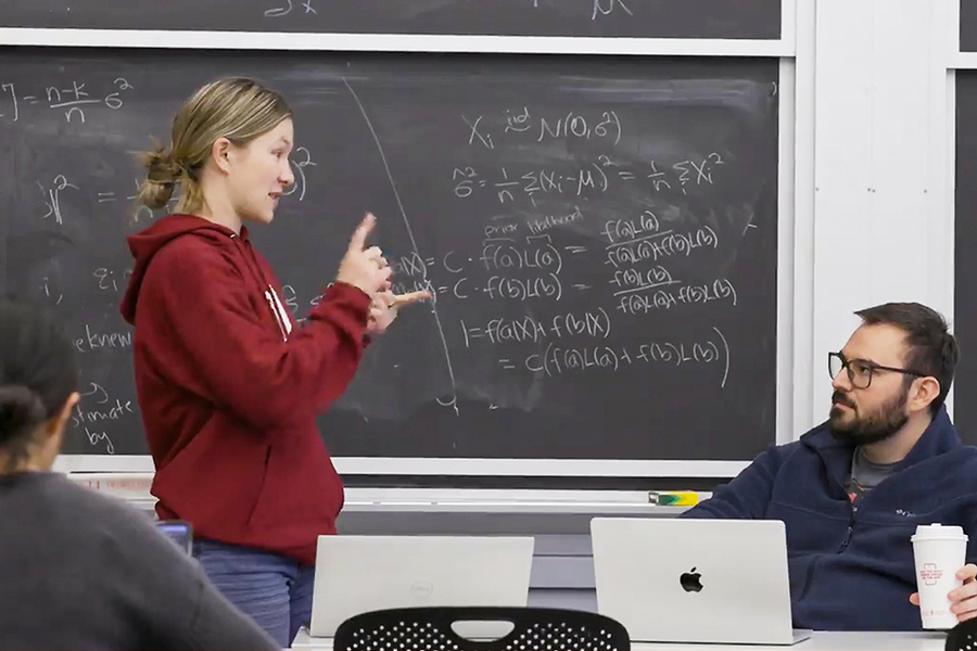 Student with ponytail speaks with instructor in a classroom with chalkboard behind them