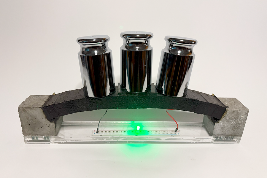 Photo of a miniature concrete arch: Rectangular, black electrodes form an arch between two concrete blocks, with metal weights on top of the arch. The system is connected to and powers a small green LED.