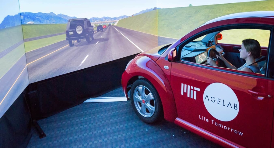 A woman steers a red coupe bearing the MIT AgeLab logo through a highway scene projected on three panels
