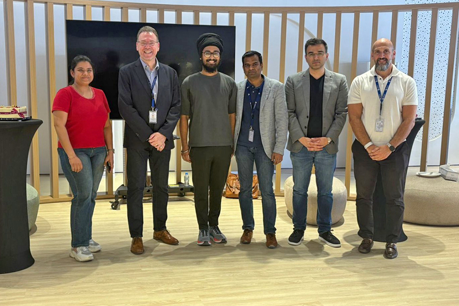 Smruthi Kilari, Andrew Murphy, Amitoj Singh, Satish Kumar, Rajeet Sampat, and Wesam Omar pose standing indoors with a blank TV screen behind them.