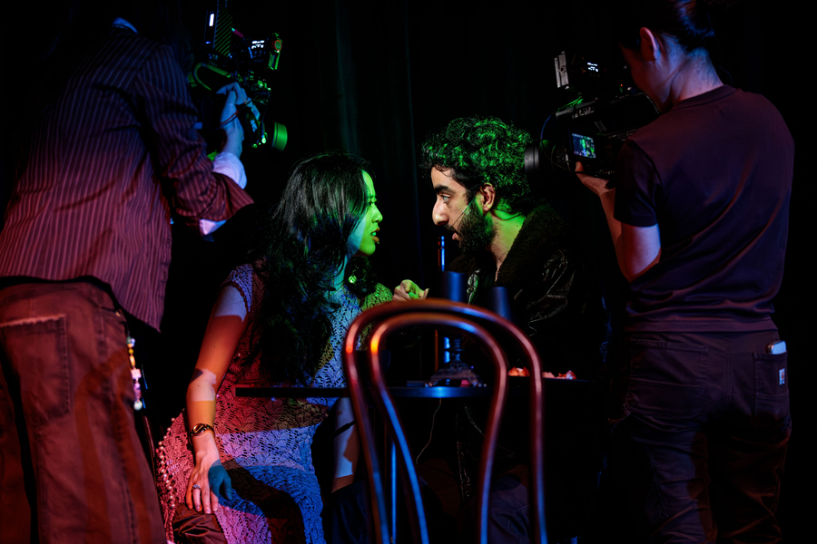 Two actors seated at a table on stage during a performance, bathed in colored lights, with camera operators filming them